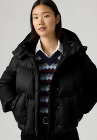 Jonge vrouw met een zwarte pufferjack over een witte blouse en een multikleurige trui met ruitpatroon, staand tegen een neutrale achtergrond.