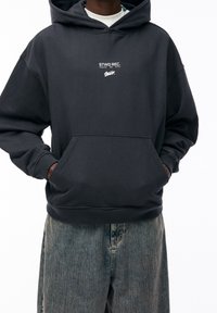 Czarna bluza z kapturem wykonana z bawełnianej mieszanki, posiadająca przednią kieszeń, ściągacze na rękawach oraz biały nadruk logo na piersi. Noszona z jasnoniebieskimi, lekko wyblakłymi, szerokimi jeansami.