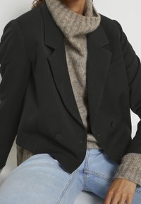 Musta kaksirivinen bleiseri huippuilevilla lapelilla, yhdistetty beigeen ribattuun pooloneuleeseen. Vaaleansiniset farkut näkyvät alaosasta.