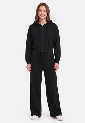 Vrouw staat glimlachend, draagt een zwarte hoodie met ritssluiting en bijpassende wijde broek met witte sneakers op een effen achtergrond.