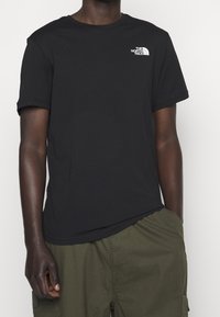 Hombre vestido con una camiseta negra de The North Face y pantalones cortos de cargo verde olivo, de pie con una mano en el bolsillo frente a un fondo blanco.