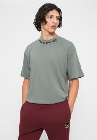 Mlad moški z temnimi kodrastimi lasmi nosi svetlo sivo majico Nike in bordo Nike trenirke, stoji z rokami v žepih in gleda desno.