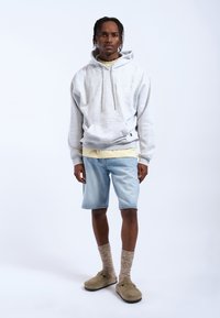 Lichtgrijze katoenen hoodie met een trekkoord en een voorkean, gelaagd over een gele T-shirt, gecombineerd met lichtblauwe denim shorts en beige instappers.