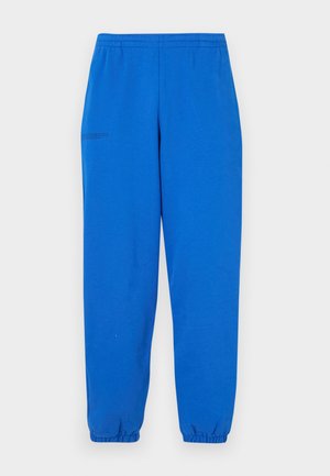 Pantalon de survêtement bleu avec une taille élastique et des poignets élastiques. Tissu lisse avec un léger éclat. Présente un petit détail imprimé sur le côté droit.