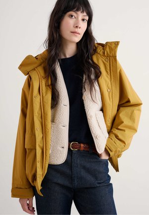 Goldene Jacke mit einem strukturierten, gefütterten Innenraum. Schwarzer Pullover darunter, kombiniert mit dunkelblauen Jeans und einem geflochtenen braunen Gürtel.
