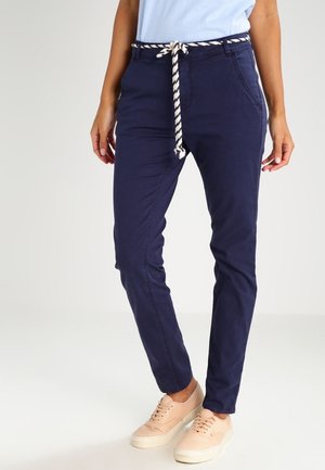 Pantalones ajustados azul marino con cintura ajustable a rayas, bolsillos frontales y detalles cosidos. Combinados con zapatillas de color claro.