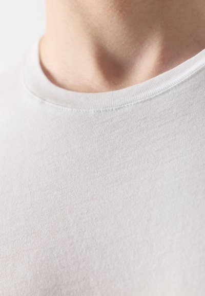Gros plan sur le cou et le haut de la poitrine d'une personne portant un t-shirt blanc uni à col ras du cou sur un fond blanc.