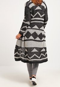 Pletený kardigan v černé a bílé barvě, s geometrickými vzory a dlouhým, otevřeným designem. Spárován se šedými legínami a bílými teniskami.