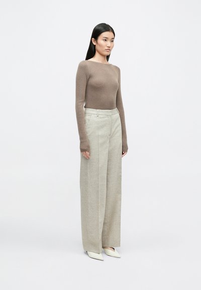 Smeđa pletena majica dugih rukava u kombinaciji sa svijetlosivim širokim hlačama i svijetlim cipelama s šiljastim vrhom, prikazana na jednobojnoj pozadini.