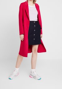 Fuchsia lång kappa över en vit och lila randig skjorta, kombinerad med en marinblå kjol med silverknappar och vita sneakers med rosa detaljer.