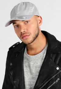 Sølv baseball cap med en blank finish, afrundet krone og buet skygge, parret med en sort læderjakke og grå skjorte.
