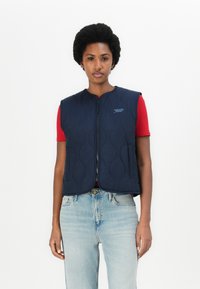 Marinblå vadderad väst med rund halsringning, dragkedjestängning och sidfickor. Bärs över en röd skjorta och ljus denimjeans.