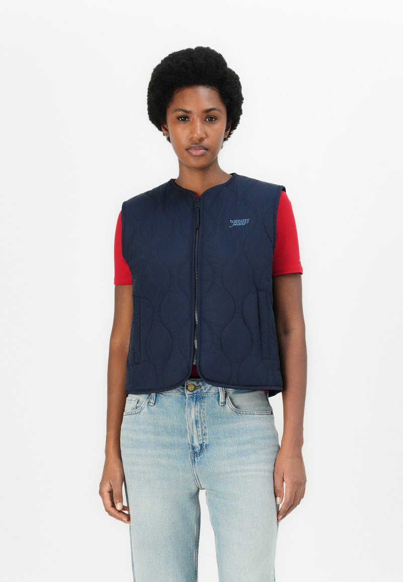 Marinblå vadderad väst med rund halsringning, dragkedjestängning och sidfickor. Bärs över en röd skjorta och ljus denimjeans.