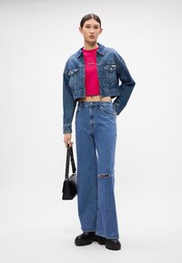 Jaqueta de ganga com um design curto, combinada com uma camisa rosa de mangas curtas, jeans rasgados de perna larga e botas de plataforma pretas. Inclui uma mala preta.