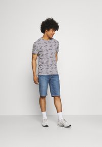 Camiseta gris con estampado de palmeras negras, de mangas cortas, combinada con pantalones cortos de mezclilla azul. El modelo lleva zapatillas blancas y calcetines por el tobillo.