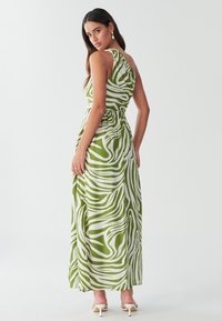 Maxikjole med grønt og hvidt virvlende mønster, enkeltskulderdesign, tætsiddende talje og flydende tekstur i stoffet. Hvide sandaler med hæl.