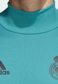 Turkusowa sportowa bluzka z długim rękawem z wysokim kołnierzem. Zawiera czarne logo Adidas oraz emblemat Real Madrid na klatce piersiowej. Gładka faktura.