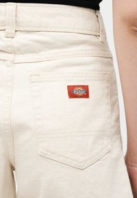 Gros plan sur la poche arrière d'un pantalon en denim blanc cassé avec une étiquette de marque Dickies rouge sur la hanche droite.