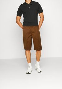 Černé polo tričko se dvěma knoflíky, ve spojení s hnědými kraťasy do kolen. Bílé sportovní boty a ponožky dotvářejí celkový vzhled.