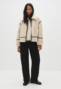 Donna con lunghi capelli scuri che indossa una giacca in shearling beige, un maglione crema, pantaloni neri a gamba larga e mocassini neri, in piedi con le mani rilassate.