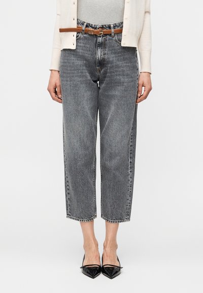 Femme portant un jean gris raccourci, un cardigan blanc, une ceinture marron et des escarpins noirs à bout pointu, debout devant un fond uni.