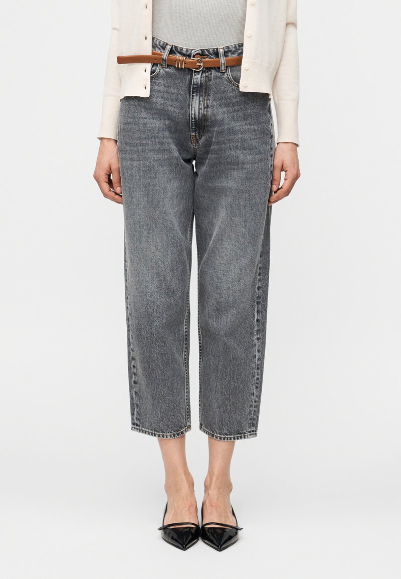 Γυναίκα με κοντά γκρι τζιν, λευκή ζ ακέτα, καφέ ζώνη και μαύρες ψηλοτάκουνες μυτερές γόβες, που στέκεται μπροστά σε μία απλή φόντο.