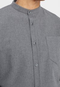 Camicia grigia con colletto alla mandarino, caratterizzata da una tasca frontale e bottoni a contrasto. Il tessuto sembra tessuto e testurizzato.