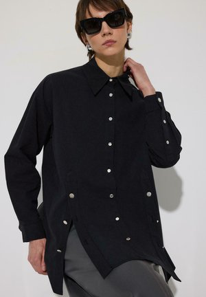 Oseba, oblečena v preveliko črno srajco z gumbi in velike črne sončne očala, ki pozira z eno roko ob vratu na enobarvnem ozadju.