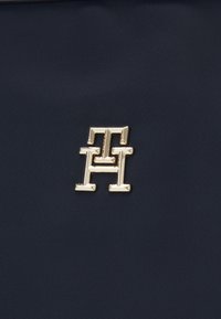 Tessuto blu marine con finitura testurizzata presenta un medaglione con logo in tono oro. Il design include forme letterarie geometriche audaci.