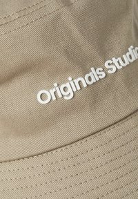 Tan bucket hat made of textured fabric, featuring a white "Originals Studio" logo. Stitching visible along the brim.