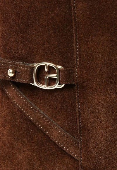 Bota de ante marrón con una hebilla plateada, con costuras contrastantes y una textura suave. Detalle notable incluye una correa decorativa.