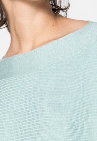Hellblau strukturierter Stoff mit einem weitem Ausschnitt und gerippten Details um den Kragen, der ein weiches, dezentes Gewebe-Muster zeigt.