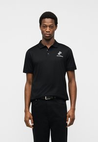 Νεαρός άνδρας που στέκεται φορώντας μαύρο πόλο μπλουζάκι με λευκό λογότυπο φοίνικα και κείμενο στο αριστερό στήθος, σε απλό γκρι φόντο.