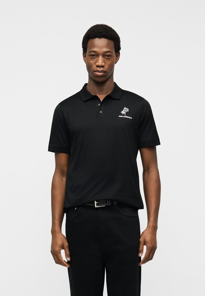 Νεαρός άνδρας που στέκεται φορώντας μαύρο πόλο μπλουζάκι με λευκό λογότυπο φοίνικα και κείμενο στο αριστερό στήθος, σε απλό γκρι φόντο.