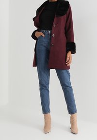 Abrigo burdeos con un collar de piel sintética negra, que cuenta con cuatro botones, llevado sobre una blusa negra y jeans azules, complementado con botas de tobillo beige.