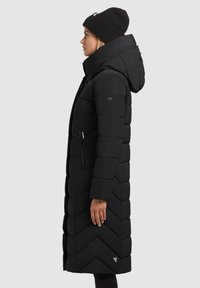 Long black puffer coat with a hood, featuring a chevron quilted design and zippered side pocket. Accompanied by a black knit beanie.