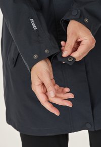 Marine waterdichte jas met drukknoopsluitingen en een gestructureerde afwerking. Voorzien van een verstelbare manchette met een metalen drukknoop en een merkdetail op de mouw.