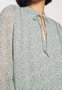 Ljusgrön blus med ett subtilt prickmönster, nyckelhålsringning med knytning och lätt puffade ärmar, tillverkad av ett texturerat tyg.