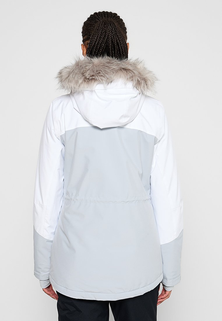 Eine Frau mit einem geflochtenen Haar, die eine hellgraue und weiße Winterjacke mit einer Kapuze aus Kunstfell trägt, steht mit dem Rücken zu einem schlichten Hintergrund.