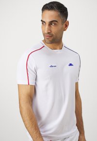 Valge sportlik T-särk, millel on punased ja sinised aktsenttriibud, tekstureeritud kangas, ümara kaelusega ning logo vasakul rindkere piirkonnas.