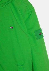 Vihreä takki, jossa kirjailtu logo ja hihassa oleva merkki. Materiaali vaikuttaa kevyeltä ja sileäpintaiselta.