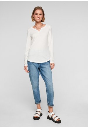 Weißes Langarmoberteil mit V-Ausschnitt, kombiniert mit hellblauen knöchellangen Jeans und weißen Sandalen mit schwarzen Sohlen und Schnallenakzenten.