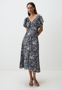 Bloemendessine, marineblauw jurkje met korte mouwen en een V-hals, gemaakt van lichte stof, vloeiend tot halverwege het scheenbeen. Zilveren sandalen.