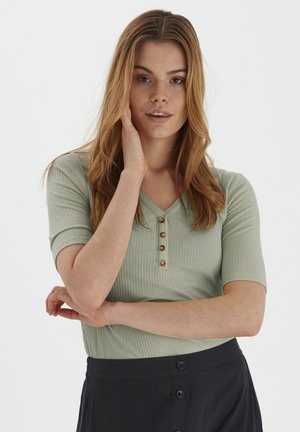 Light green ribbed top with a V-neck and short sleeves, featuring five decorative buttons on the front. Worn with a black skirt.