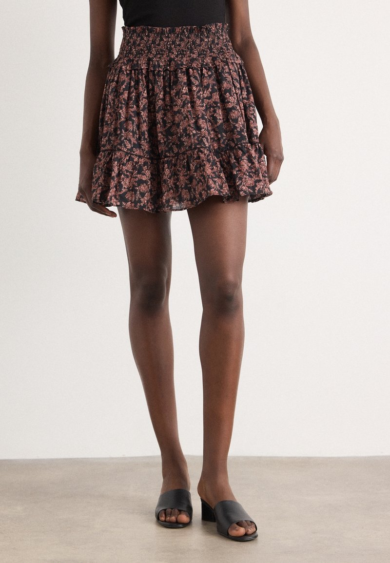 Femme portant une mini-jupe noire et rose à fleurs avec volants, associée à des sandales noires à talons blocs à bout ouvert sur un fond neutre.