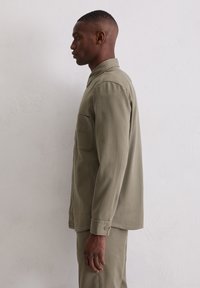 Hellolivgrünes Hemd mit Knopfleiste, Kragen, Seitentasche und langen Ärmeln. Hergestellt aus einem glatten Stoff mit lockerer Passform.