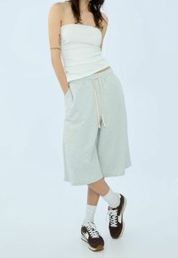 Světlé šedé teplákové kraťasy ke kolenům s elastickým pasem a šňůrkou, spárované s bílým přiléhavým tílkem a hnědými teniskami.