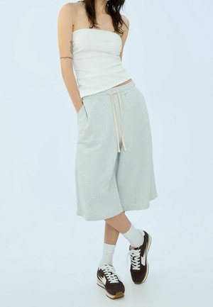 Shorts de sweat gris clair, arrivant aux genoux, avec une taille élastique et un cordon de serrage, associés à un débardeur blanc ajusté et des baskets brunes.