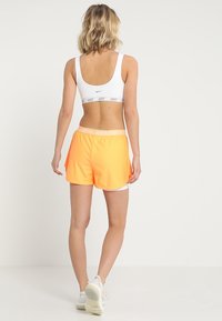 Sostenedor deportivo blanco con un logo gris de Nike, combinado con pantalones cortos de un brillante color naranja que cuentan con una cinturilla blanca de Under Armour. Textura suave, ajuste atlético.
