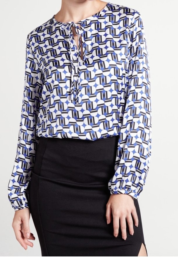 Blusa a maniche lunghe bianca con motivo geometrico blu e nero, caratterizzata da un collo con fiocco e tessuto morbido. Abbinata a una gonna nera.